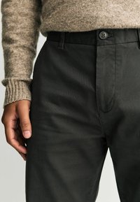 Fekete nadrág sima anyagból, elülső gombos zárással és övhurkokkal, részben látható szürke pulóverrel a tetején.