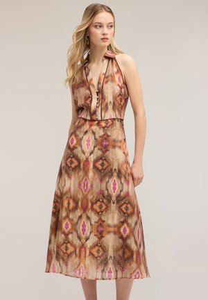 Vrouw met een mouwloze, met knopen gesloten jurk met kraag en bruine, oranje en roze geometrische patronen, staand tegen een beige achtergrond.