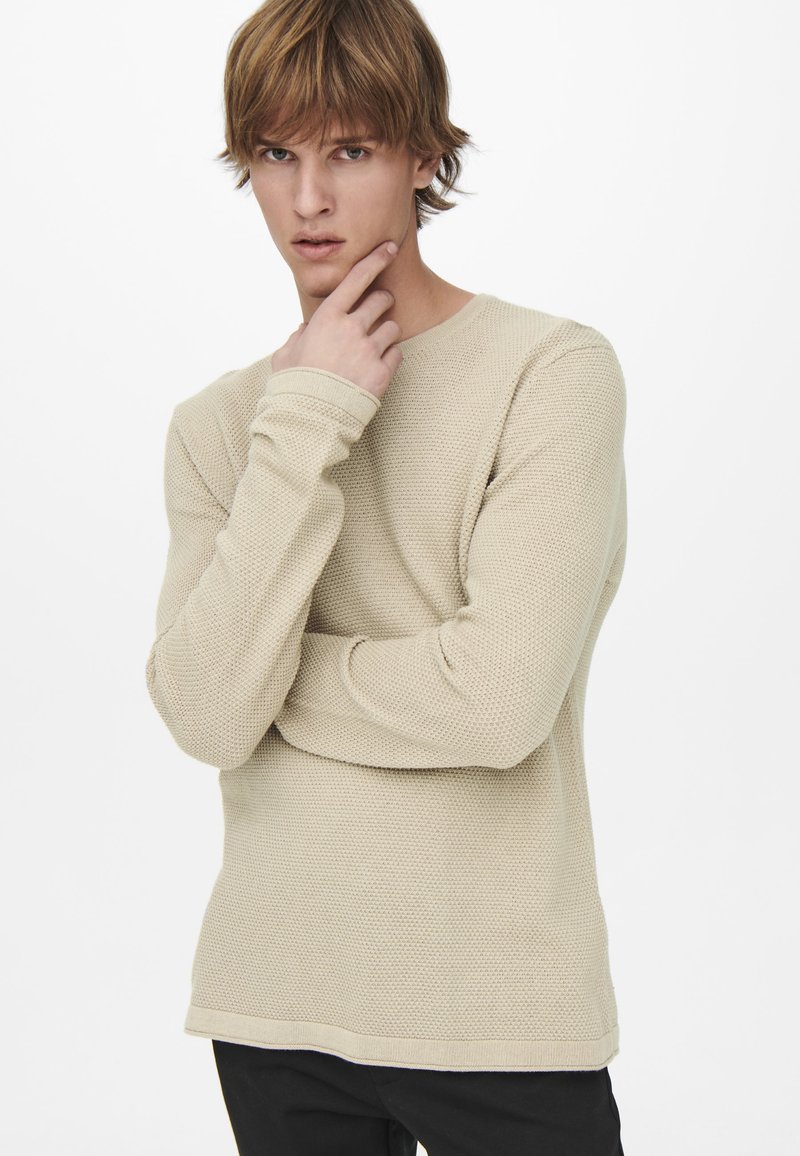 Suéter de punto texturizado color beige con mangas largas y cuello redondeado, combinado con pantalones negros. La modelo adopta una pose pensativa.