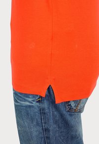 Kortärmad orange skjorta med splittad fåll, i kombination med blå denimjeans. Tyget verkar mjukt med en slät yta.