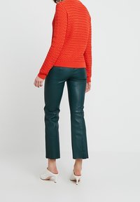 Pull en tricot rouge avec un motif texturé, assorti à un pantalon en cuir vert ajusté et des talons ouverts blancs. Accent mis sur les détails de la tenue.