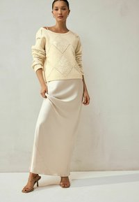 Strickpullover in Crème mit Rautenmuster, kombiniert mit einem seidigen, beigefarbenen Maxirock. Modell trägt transparente Riemchenhighheels und steht vor einem hellen Hintergrund.