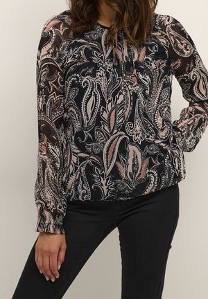 Woman wearing a long-sleeve, black paisley print blouse with sheer sleeves and black pants, standing against a plain light wall.