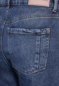Jeans de mezclilla en azul medio con una textura ligeramente desgastada. Presenta un bolsillo trasero con costuras en contraste y una etiqueta de cuero arriba.