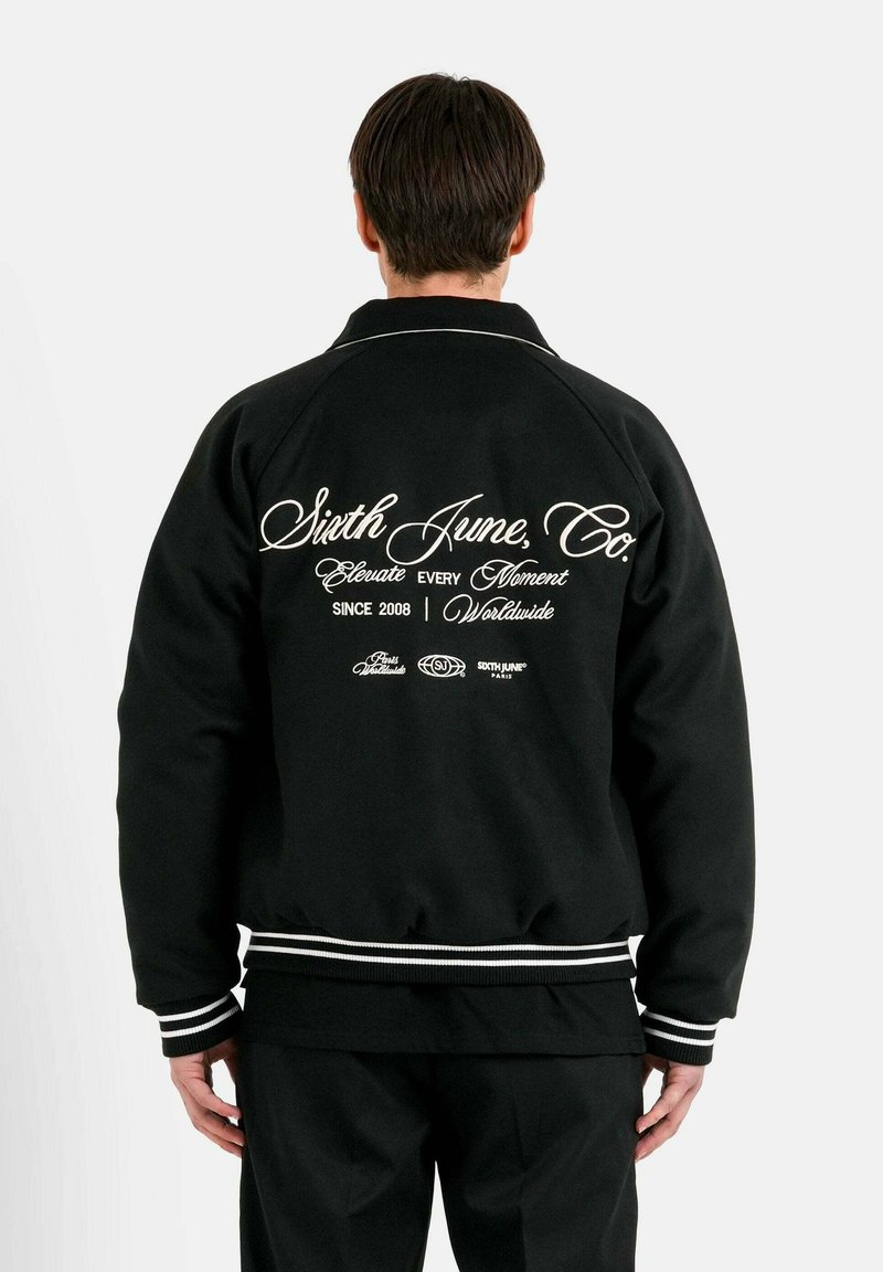 Schwarze Jacke mit weißen Akzenten, ausgestattet mit einem gerippten Kragen und Bündchen. Großes grafisches Textdesign auf der Rückseite, einschließlich Markenname und Slogan.