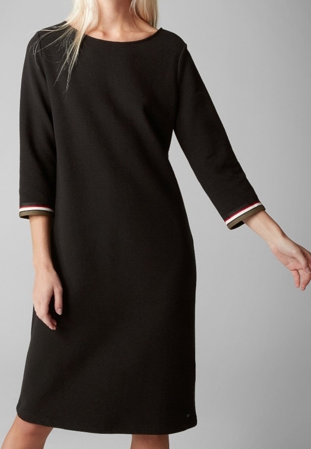 Femme portant une robe noire longueur genou avec des manches trois-quarts ornées de poignets rayés rouge, blanc et beige, sur un fond gris.