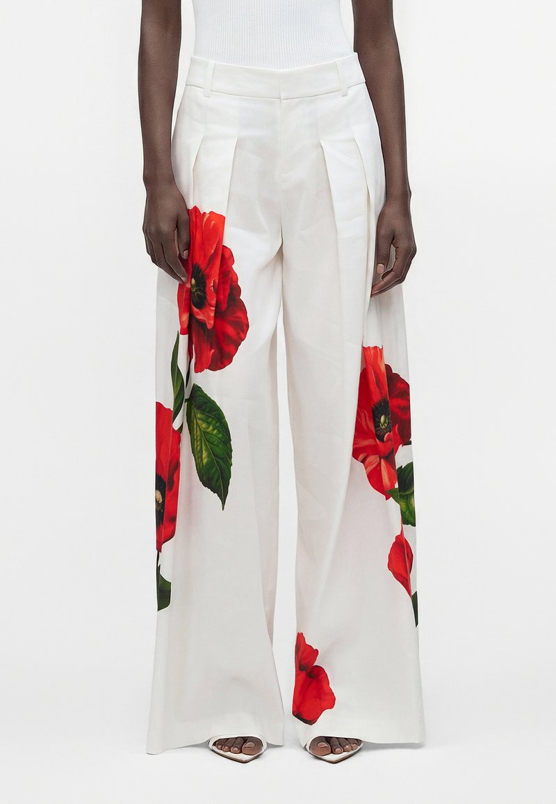 Pantalon blanc à jambes larges avec de grands imprimés de coquelicots rouges et de feuilles vertes, porté avec des sandales blanches à talons.