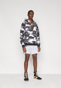 Φούτερ με κουκούλα σε ασπρόμαυρο σχέδιο και πορτοκαλί κορδόνια, συνδυασμένο με λευκά σορτς και μαύρα αθλητικά παπούτσια. Διαθέτει ριμπ τελείωμα και λεπτομέρειες με λογότυπο.