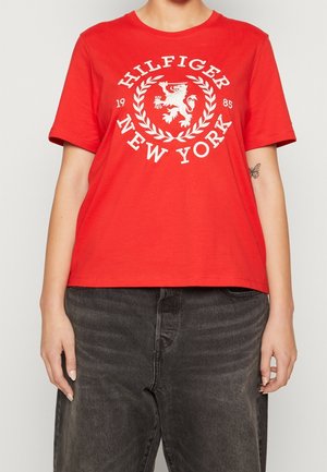 Person wearing a red short-sleeve shirt with "Hilfiger New York 1985" white logo and faded black jeans, standing with arms relaxed.