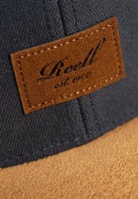 Dunkelblaues Stoffmütze mit einem braunen Lederpatch, das den Markennamen und das Gründungsdatum zeigt, kontrastiert mit einer strukturierten beigen Unterseite.