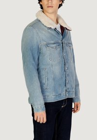 Giacca di jeans azzurro con colletto sherpa, chiusura con bottoni, due tasche sul petto e tasche laterali. Indossata sopra una camicia scura.