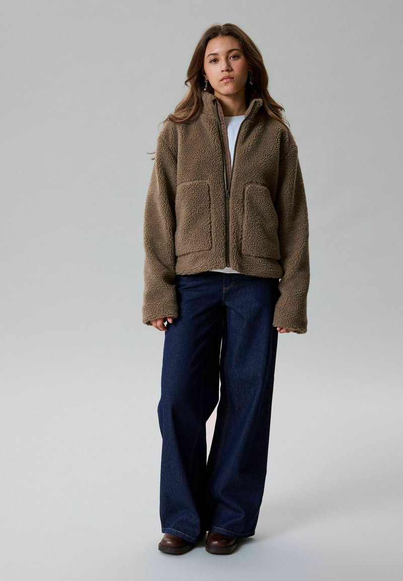 Jeune femme portant une veste polaire marron, un haut blanc, un jean bleu foncé à jambes larges et des chaussures marron, debout devant un fond gris uni.