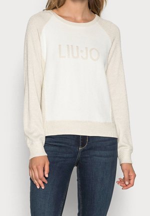 Vrouw draagt een crèmekleurige trui met lange mouwen en de tekst "LIU JO" op de voorkant, gecombineerd met donkerblauwe spijkerbroek.