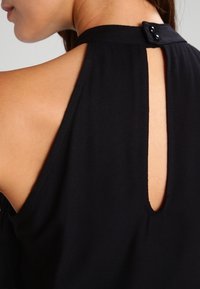 Schwarzes ärmelloses Top mit einem Schlüssel-Ausschnitt am Rücken, einer Knopfverschluss am Hals und einer glatten Textur.