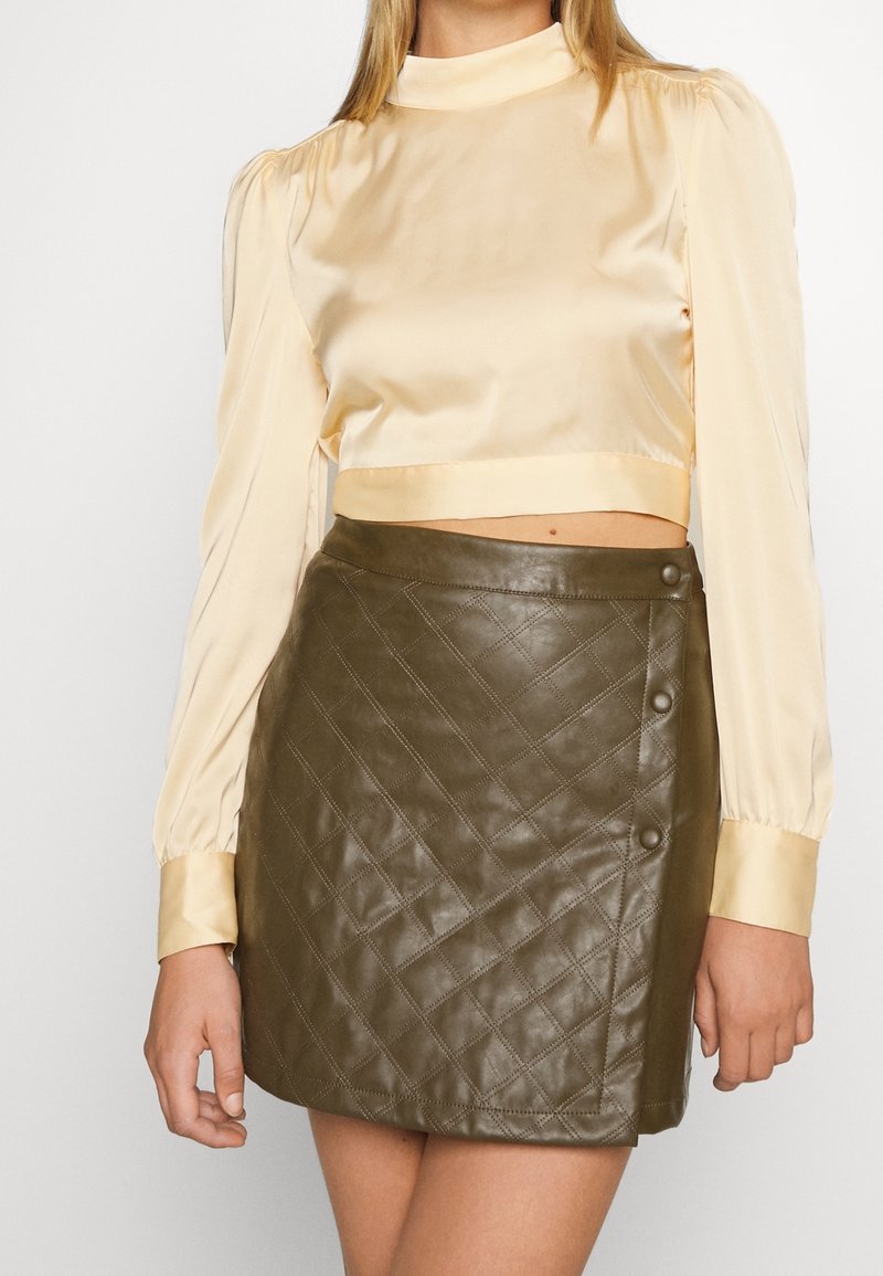 Blouse longue en satin crème à manches longues et col montant, associée à une mini-jupe en cuir matelassé vert olive avec des boutons décoratifs.