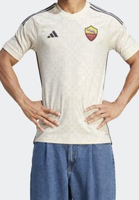 Jersey Adidas color crema que presenta un patrón sutil, acentos negros, un cuello redondo y un logo de la AS Roma en el pecho.