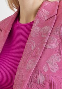 Blazer texturé rose avec motif floral placé sur un haut rose vif à côtes, mettant en valeur un tissu distinctif et un contraste de couleurs riche.