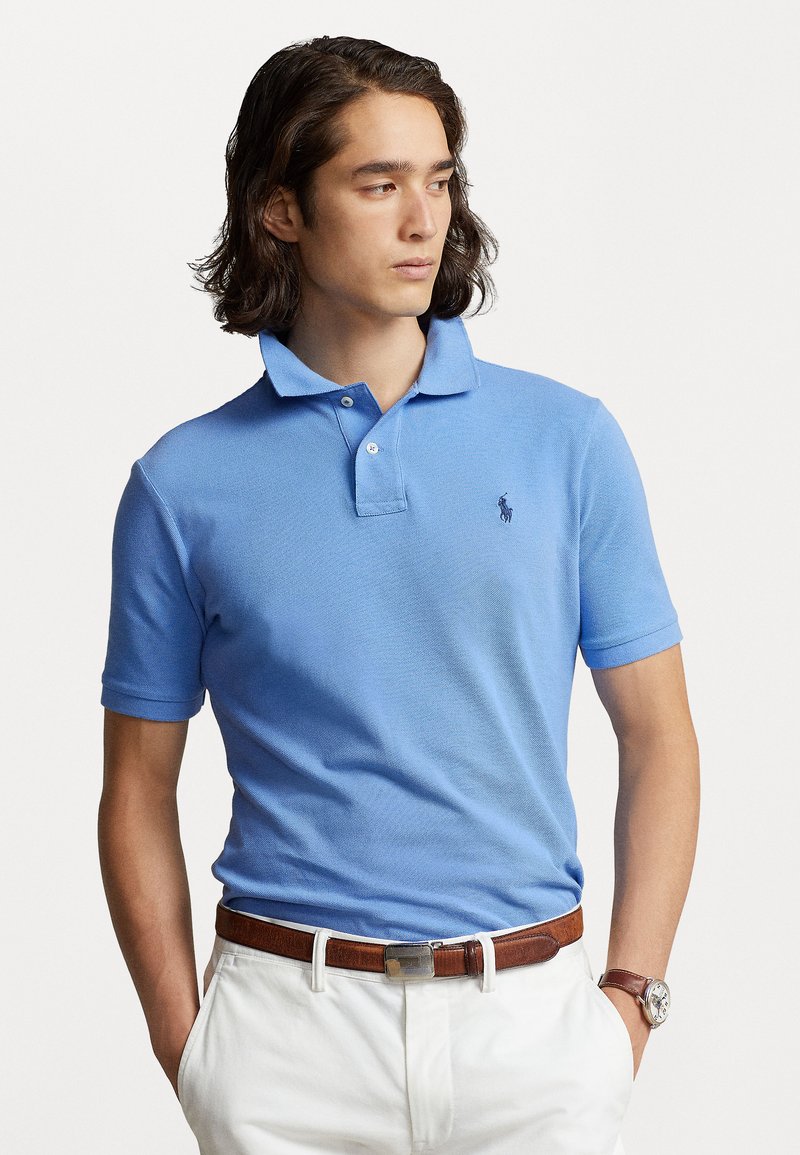Polo bleu clair avec un col, des manches courtes et un petit logo brodé. Assorti avec un pantalon blanc et une ceinture marron.