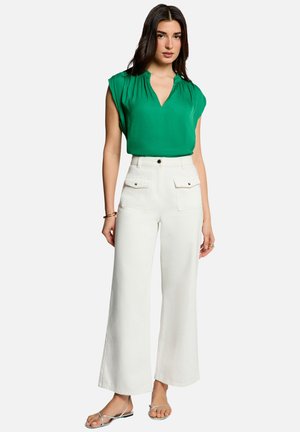 Femme portant un chemisier vert sans manches, un pantalon blanc taille haute à jambes larges avec poches avant, et des sandales argentées à brides, debout sur fond blanc.