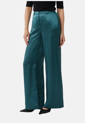 Szersze spodnie w kolorze turkusowym, wykonane z błyszczącego, teksturowanego materiału, z wysokim stanem, przednimi kieszeniami i paskiem. W połączeniu z czarnymi butami.