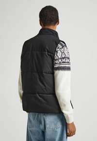 Musta pehmustettu liivi, jossa korkea kaulus ja kuvioidut valkoiset ja laivastonsiniset neulotut hihat. Selkäpuoli on sileä ja ompelut tasaiset.