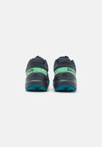 Des chaussures de sport avec une tige en maille marine, des accents verts et noirs, une semelle grise robuste et une semelle extérieure texturée bleue pour une meilleure adhérence.
