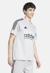 Camiseta deportiva blanca de Adidas con rayas negras, con cuello redondo, mangas cortas y logo impreso en la parte frontal. Diseño de tela texturizada.