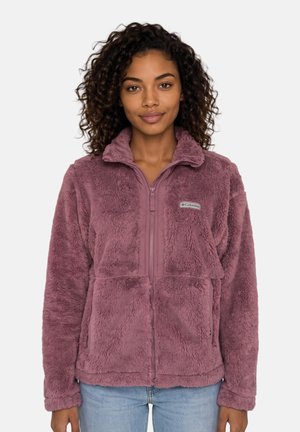 Donna con capelli ricci che indossa una giacca in pile Columbia malva con tasche frontali e cerniera, in piedi davanti a uno sfondo semplice.