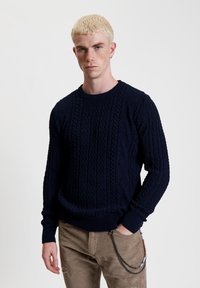 Maglione lavorato a maglia blu navy con motivi a treccia testurizzati, polsini e orlo a costine. Indossato con pantaloni in corduroy beige con un accento a catena.