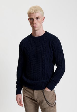 Granatowy sweter z dzianiny z teksturowymi wzorami w warkocze, z prążkowanymi mankietami i dołem. Noszony z beżowymi spodniami z kordurowej tkaniny z akcentowanym łańcuchem.