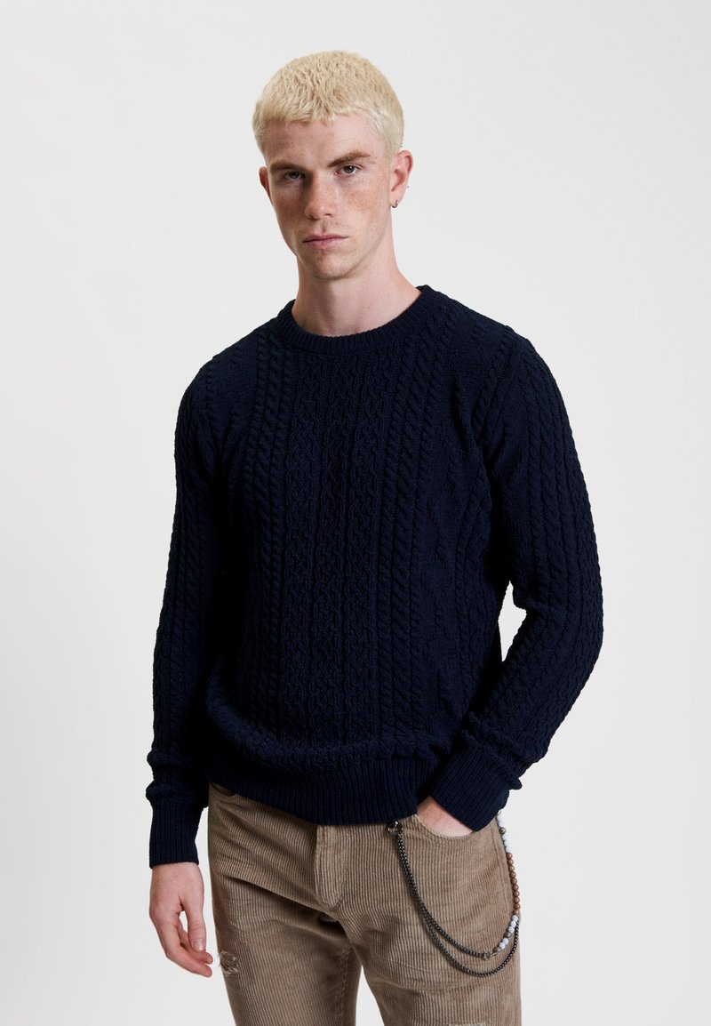 Maglione lavorato a maglia blu navy con motivi a treccia testurizzati, polsini e orlo a costine. Indossato con pantaloni in corduroy beige con un accento a catena.