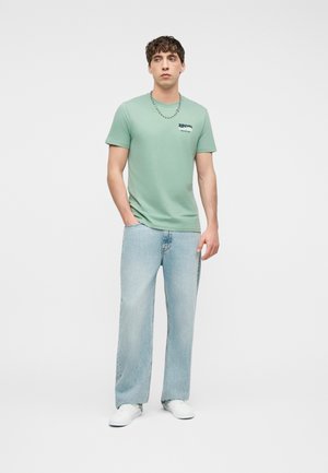 Giovane uomo in piedi che indossa una maglietta verde chiaro RIP CURL, jeans azzurri, sneakers bianche e una collana d'argento su uno sfondo semplice.