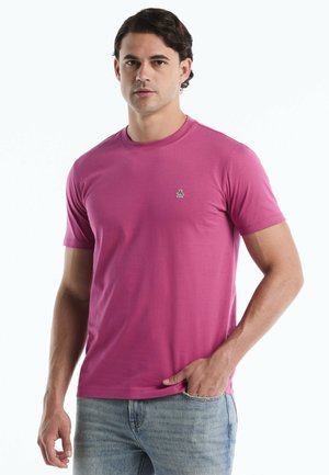 Man met kort donker haar, gekleed in een effen roze T-shirt met korte mouwen en lichtblauwe jeans, staand met één hand in zijn zak.