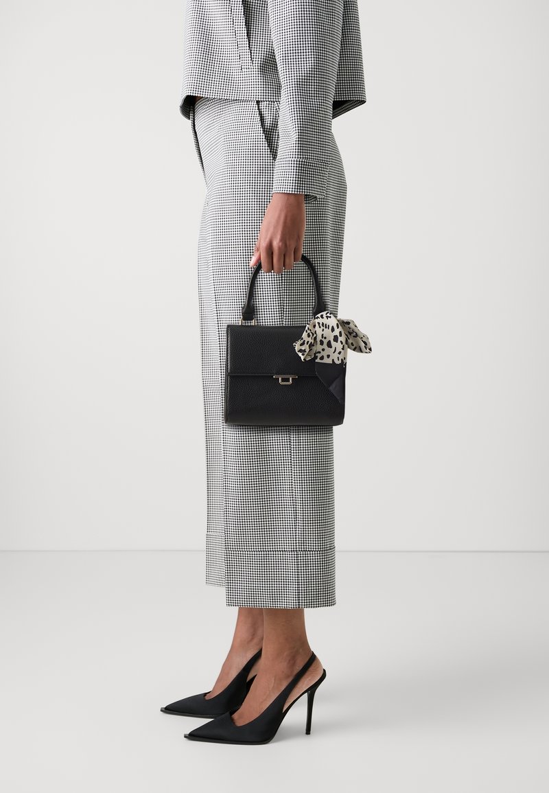 Sac à main noir texturé avec une poignée supérieure et un nœud en tissu à motif. Modèle portant une tenue grise à carreaux et des chaussures à talons noirs.