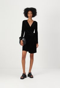 Vestido envolvente preto em veludo, com mangas longas, decote em V, detalhe de laço lateral e barra curta. Combinado com uma bolsa circular preta e sandálias chunky.