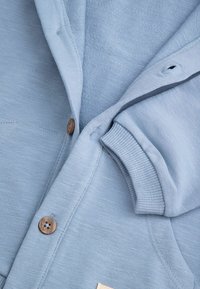 Jasnoniebieska, rozpinana bluza z dzianiny, wyróżniająca się fakturowanym materiałem, drewnianymi guzikami i ściągaczami przy mankietach. Miękki, casualowy fason z kapturem.