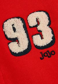 Crvena tkanina sa bijelim, pahuljastim izvezenim brojevima "93" obrubljenim crnom, uz ime "Jojo" izšiveno crnom koncem ispod brojeva.