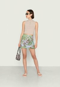 Top bege sem mangas com gola alta, saia mini estampada em verdes e azuis, bolsa preta com riscas brancas e sandálias bege.