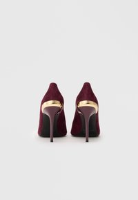 Ζευγάρι μπορντώ σουέτ γυναικείων παπουτσιών με ψηλά τακούνια και χρυσές ιμάντες με την επιγραφή "LOVE MOSCHINO" στην πίσω πλευρά.