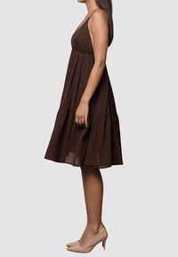 Vestido marrón de capas hecho de un tejido ligero, con tirantes finos y una silueta fluida. Combinado con zapatos de tacón altos en tono nude.