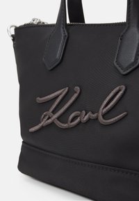 Czarna torebka z nylonu z skórzanymi akcentami, dwoma górnymi uchwytami i wyhaftowanym logo "Karl" w brązowej nici z przodu.