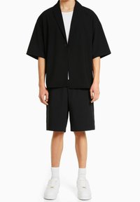 Musta ylisuuria bleiseri lyhyillä hihoilla, jossa on yksi nappi, yhdistetty mustiin shortseihin; päällä valkoinen T-paita ja valkoiset lenkkarit.