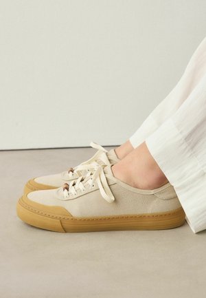 Beige canvas sneakers with light brown rubber soles and toe caps, laced up, worn with white pants, resting on a light floor against a plain wall.