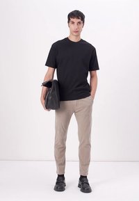 Tricou negru din bumbac, pantaloni chino bej, slim-fit, pantofi din piele negri. Modelul ține o geantă din piele neagră, stând în fața unui fundal alb simplu.