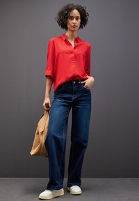 Camicia rossa con bottoni, jeans larghi blu scuro e sneakers bianche. Tiene una borsa in suede beige. Tessuto morbido e silhouette rilassata.