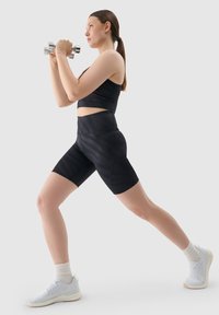 Czarny sportowy stanik i wysokie szorty z subtelnym wzorem w paski. Modelka trzyma hantle, ma na sobie białe sportowe buty i prążkowane skarpetki.