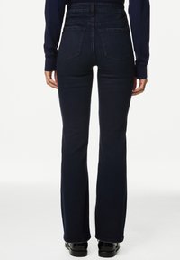 Persona vistiendo jeans de tiro alto en azul oscuro con bolsillos traseros y zapatos negros, de pie contra un fondo blanco liso.