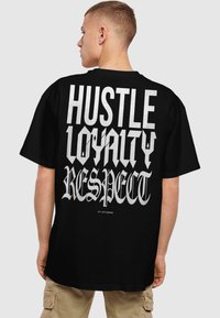 Melna kokvilnas T-krekls ar lieliem baltiem burtiem uz aizmugures: "HUSTLE LOYALTY RESPECT" dažādos treknos fonta stilus.