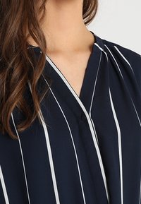 Marinblus med vita vertikala ränder, med V-ringning, knappdetaljer och mjuk, slät textur. Blusen har en avslappnad passform.