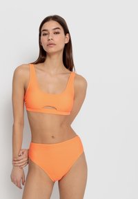 Nabor oranžne bikinkini, ki vključuje zgornji del z izrezom na sredini in hlačke z visokim pasom, izdelane iz gladkega, elastičnega materiala.